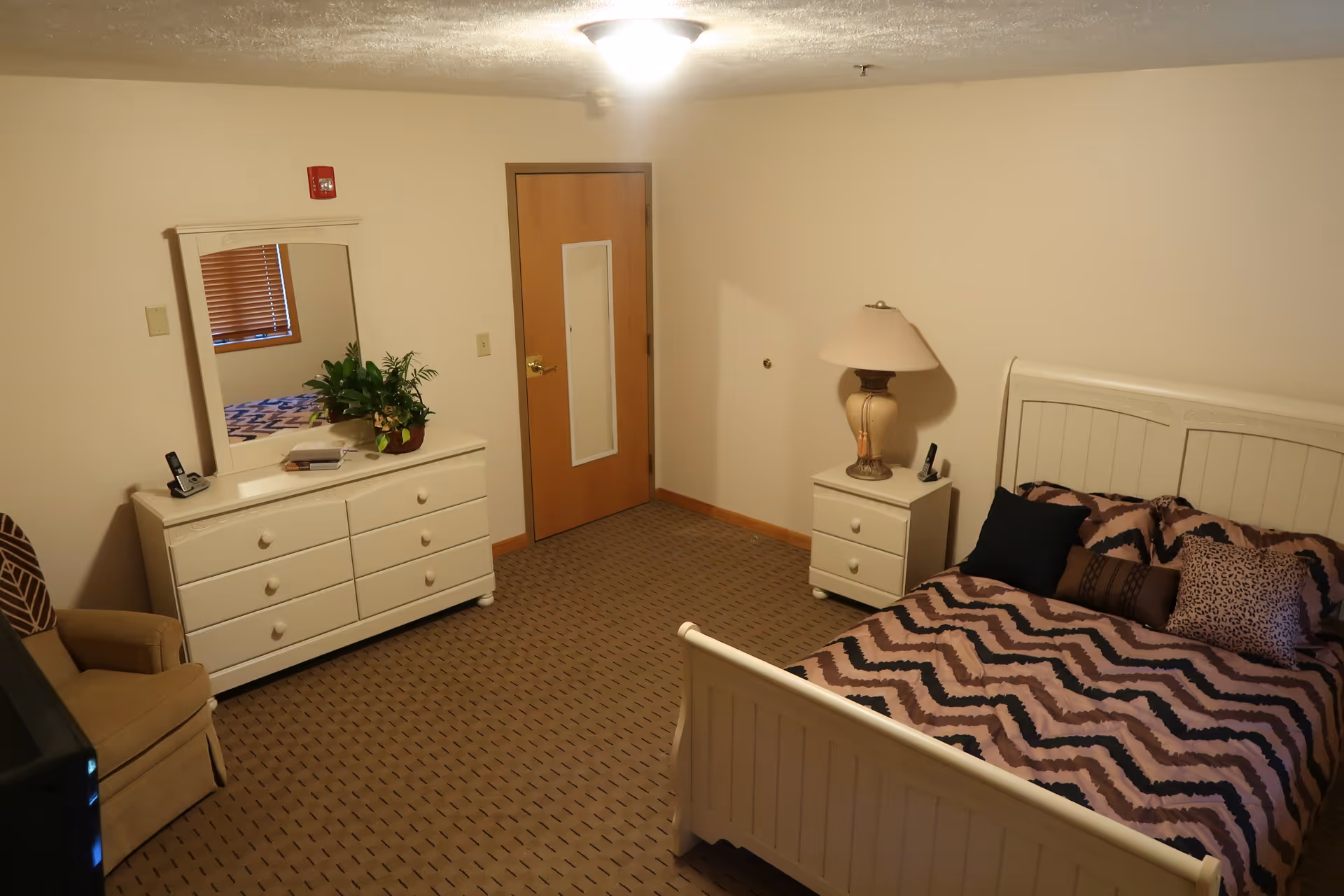 Neatly furnished bedroom with a bed, nightstand, dresser with mirror, armchair, and lamp.
