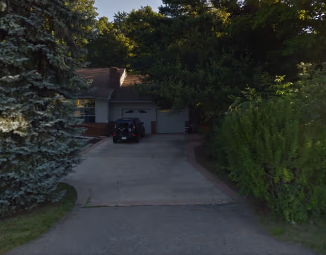 Front exterior of a single-story senior facility partially obscured by trees, with a driveway and garage.