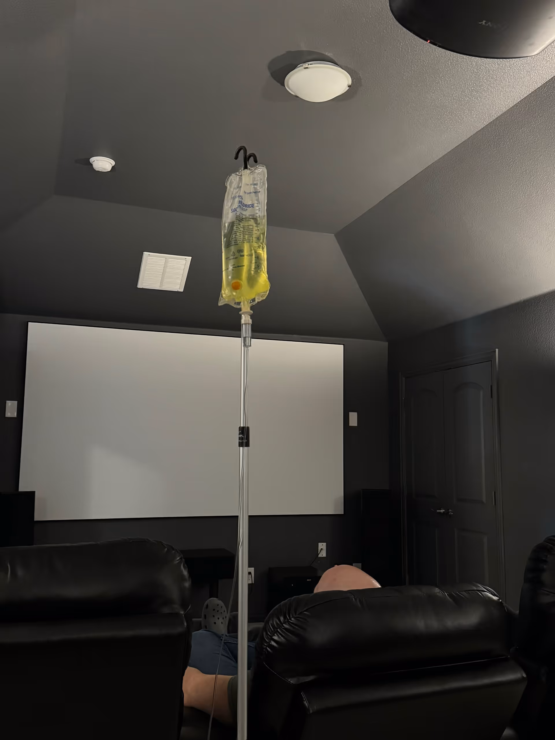 IV drip bag on stand in a dark living room with recliners and a screen.