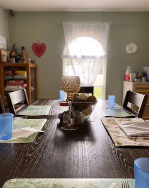 A dining table set for four with placemats, blue drinking glasses, and napkins. In the center of the table is a decorative bowl with items and a candle holder. In the background, there is a window with sheer white curtains, a wall clock, and a wooden shelf with various items including a red heart decoration.
