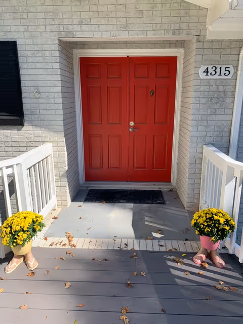 Red front door with white trim, brick facade, and potted yellow chrysanthemums on a porch.