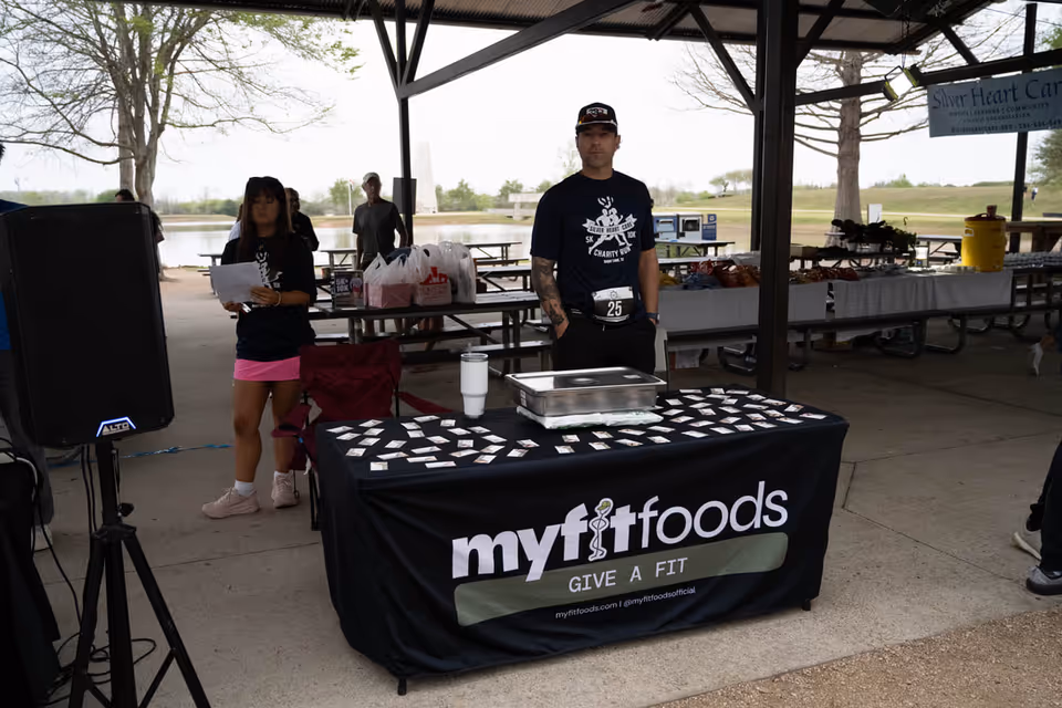 Outdoor pavilion with a table covered by a myfitfoods banner and people at an event.