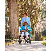 Caregiver pushes an elderly woman in a wheelchair along a sunny outdoor sidewalk.