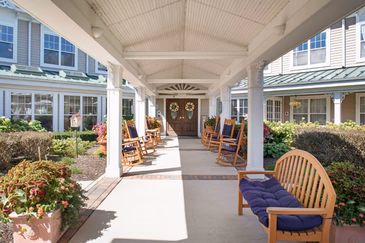 Covered front entrance walkway of a senior living facility with rocking chairs, benches, potted plants, and double doors decorated with wreaths.