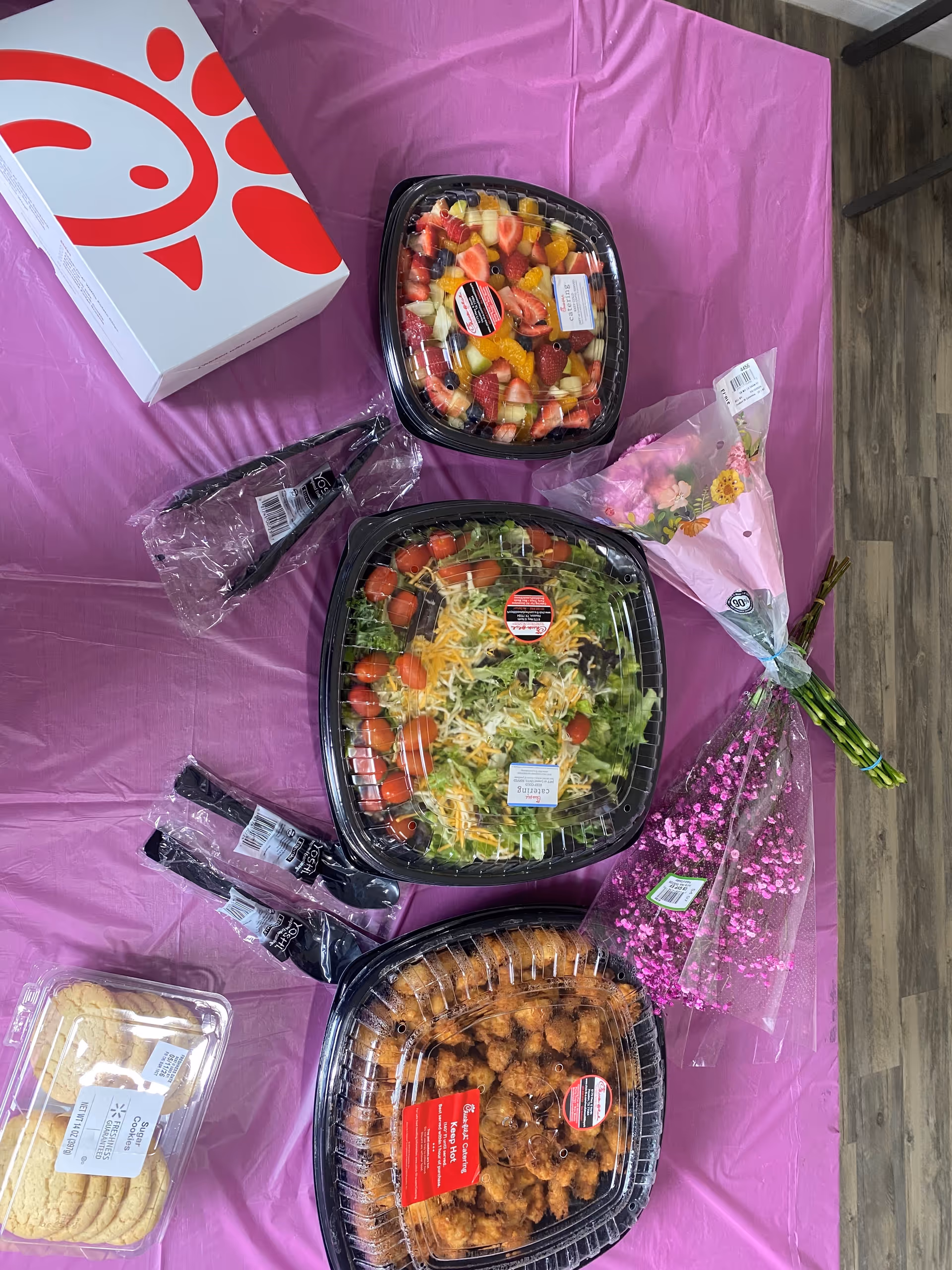 Table with pink cloth displaying fruit salad, greens salad, pastries, and flowers.