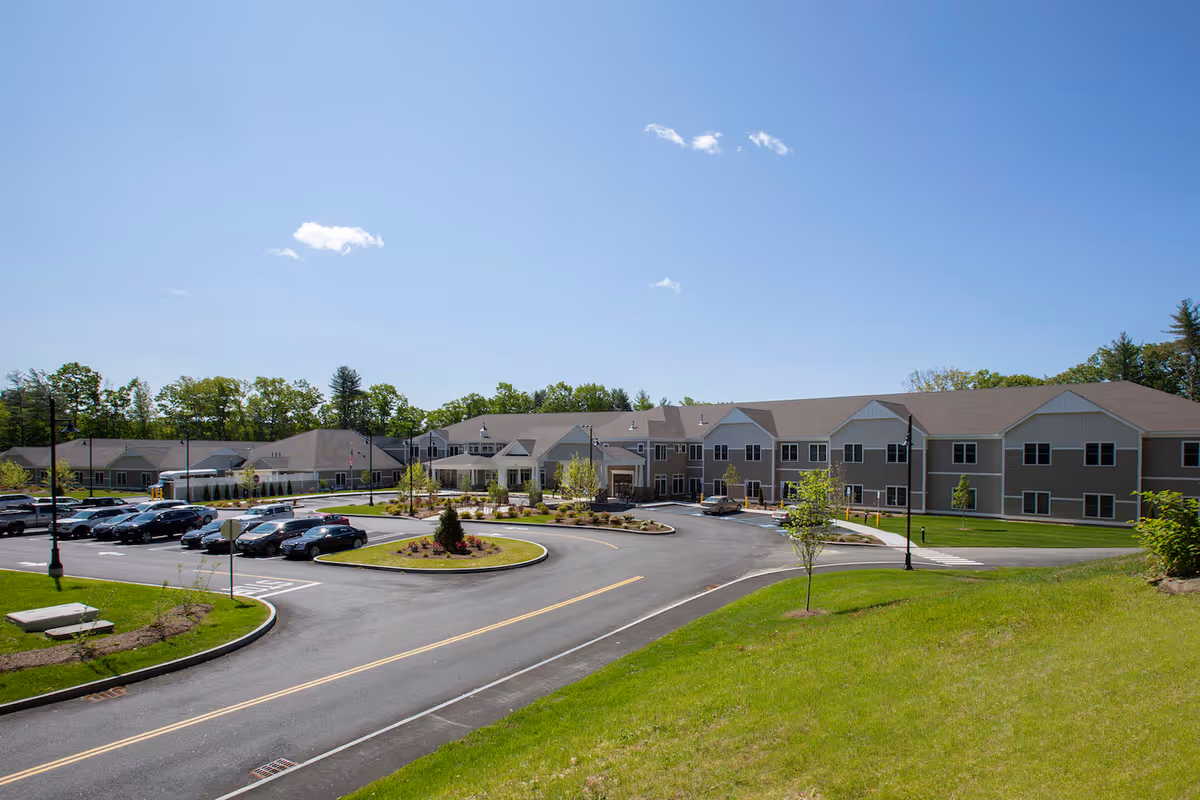 Front exterior of a two-story senior living facility with a circular driveway, parking lot, and landscaped lawn under a clear blue sky.