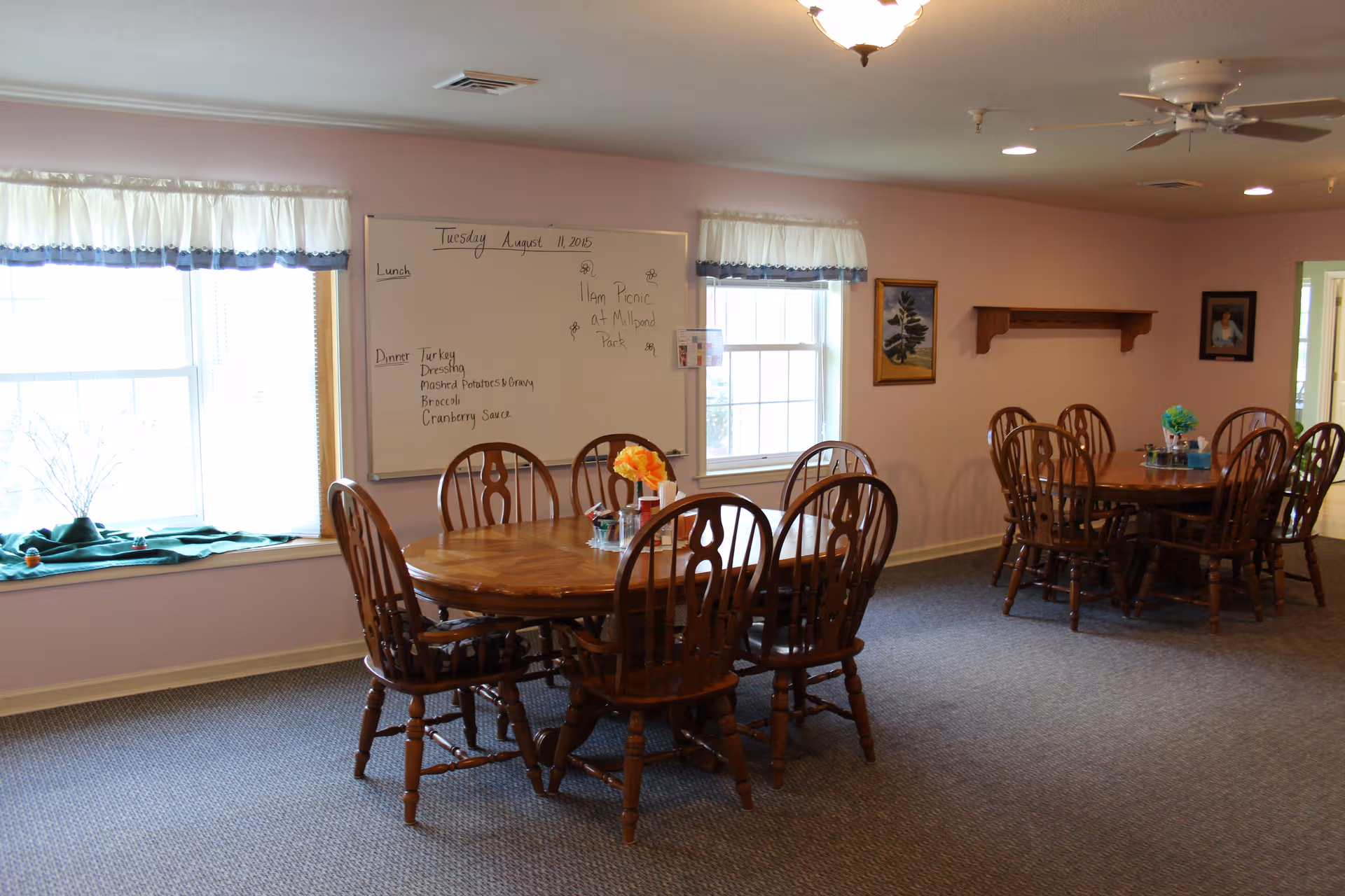 Pastel dining room with round tables, wooden chairs, windows, and a whiteboard.