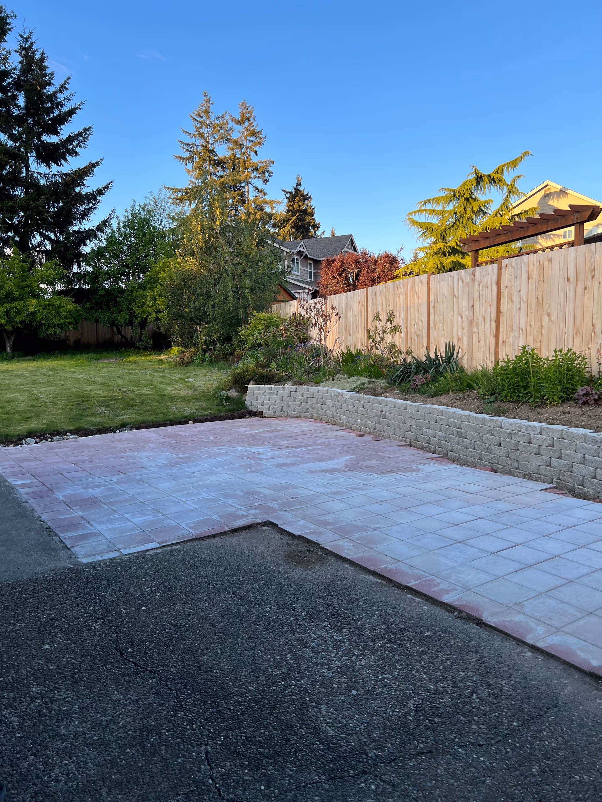Backyard patio with tiled surface, raised garden bed, wooden fence, and trees under blue sky.