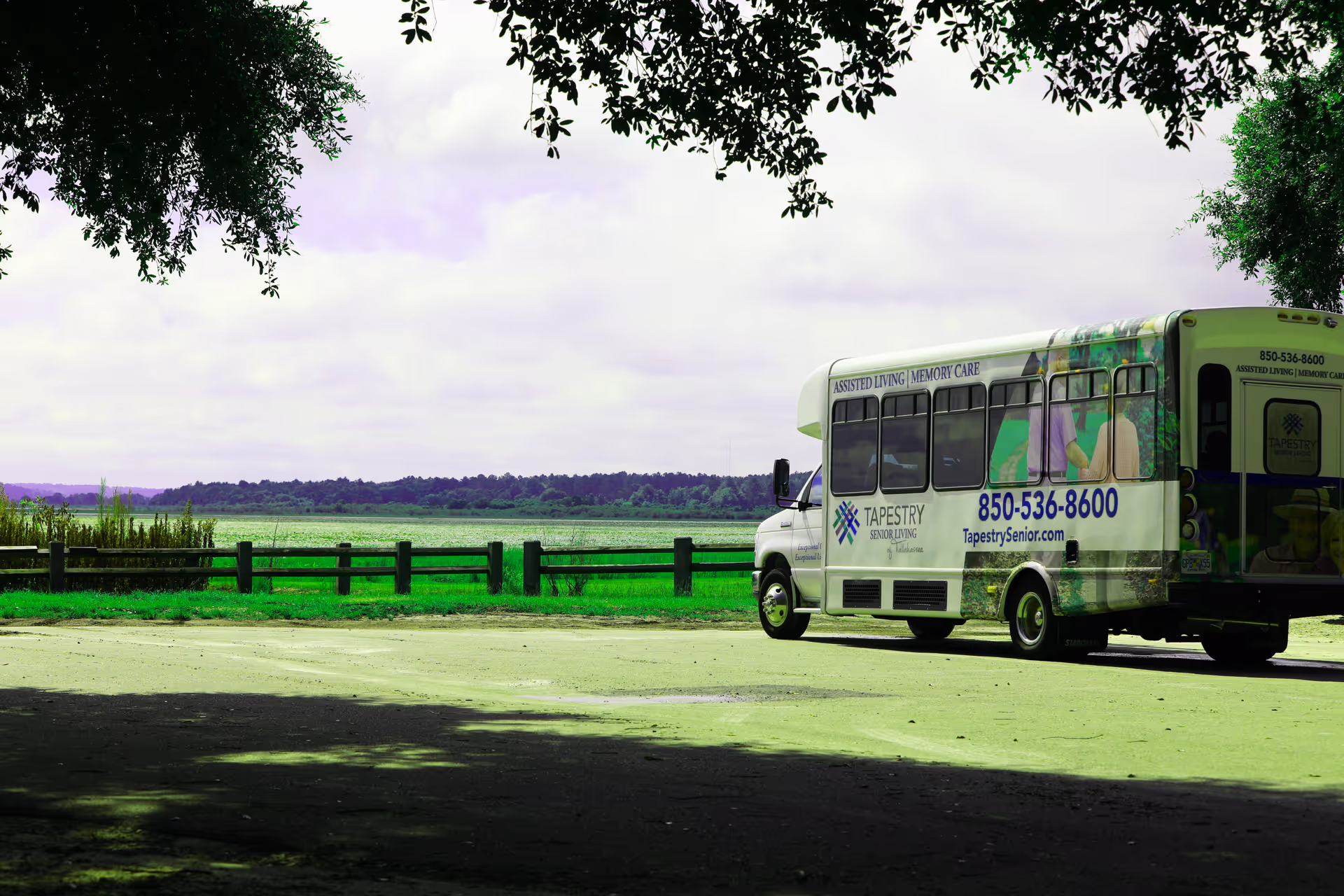 A white shuttle bus parked near a wooden fence with a scenic view of green fields and a cloudy sky in the background. The bus has signage for Tapestry Senior Living Lakeshore, including their phone number and website.