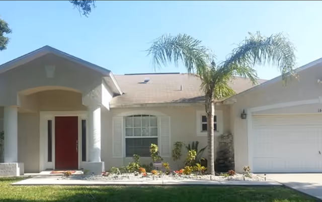 Front exterior of a single-story house with a red entry door, attached garage, palm tree, and a small flowerbed.