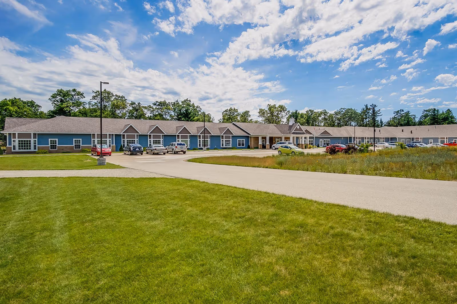 Front view of a single-story assisted living facility with a long blue building, driveway with parked cars, and a large grassy lawn under a partly cloudy sky.