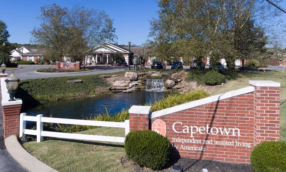 Front entrance of Capetown Senior Living showing a brick sign, pond with a small waterfall, landscaping, and the facility building in the background.