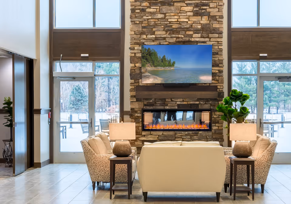A cozy seating area in a senior living facility featuring a stone fireplace with a modern electric fire and a large TV mounted above it displaying a scenic lake view. The seating includes a cream-colored sofa and two patterned armchairs, each with a side table and lamp. Large windows and glass doors on either side provide natural light and a view of outdoor seating and trees.