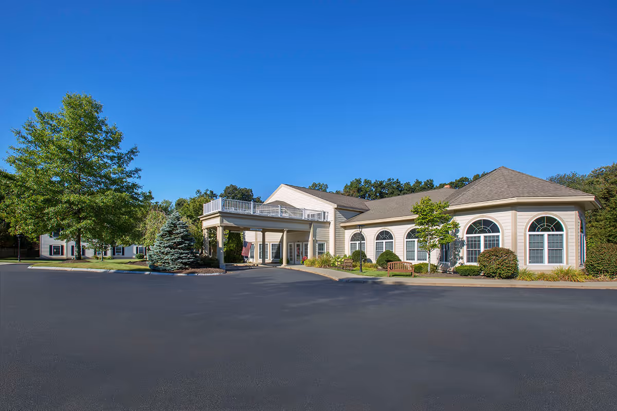 Front exterior of a single-story senior living building with an entrance portico, arched windows, landscaping and a clear blue sky.