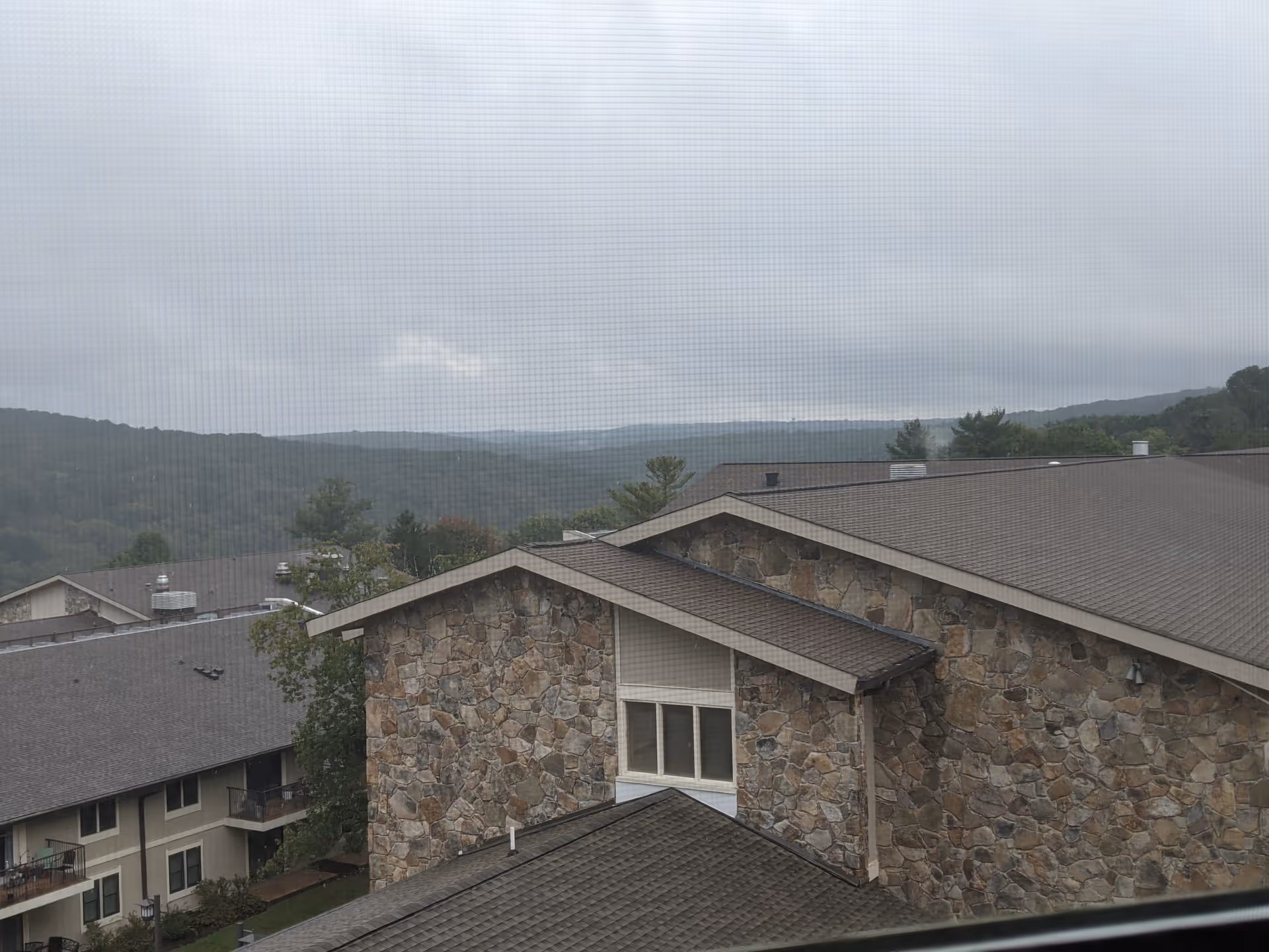 Stone-clad senior living building with sloped roofs and distant hills under a cloudy sky.