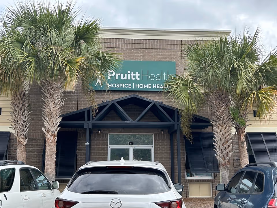 PruittHealth Hospice facility entrance with teal sign, brick facade, palm trees, and parked cars.