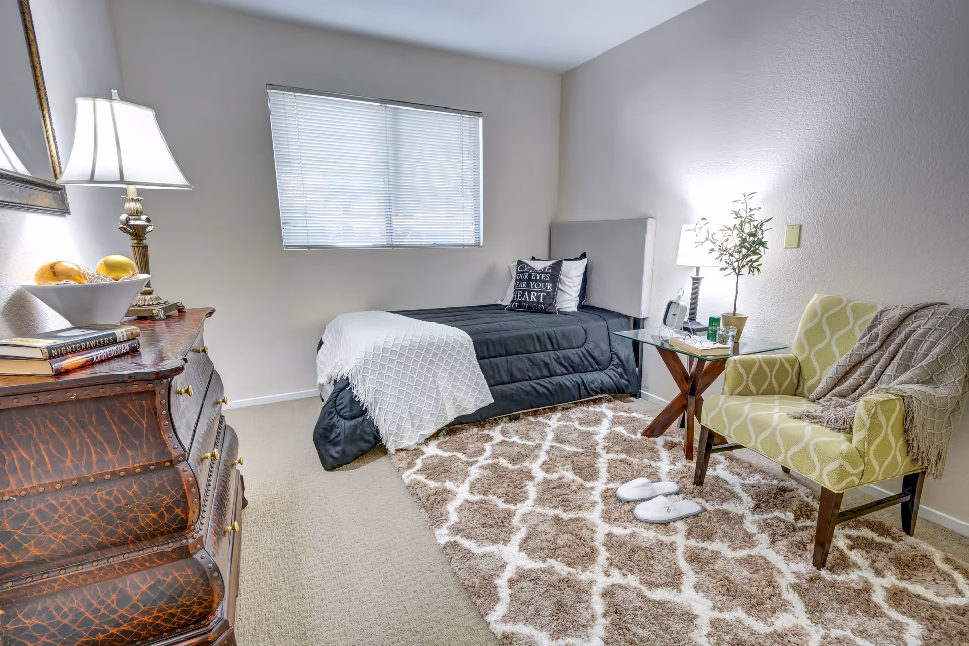 Furnished bedroom with a single bed, wooden dresser, armchair, bedside table and lamp on a patterned rug.
