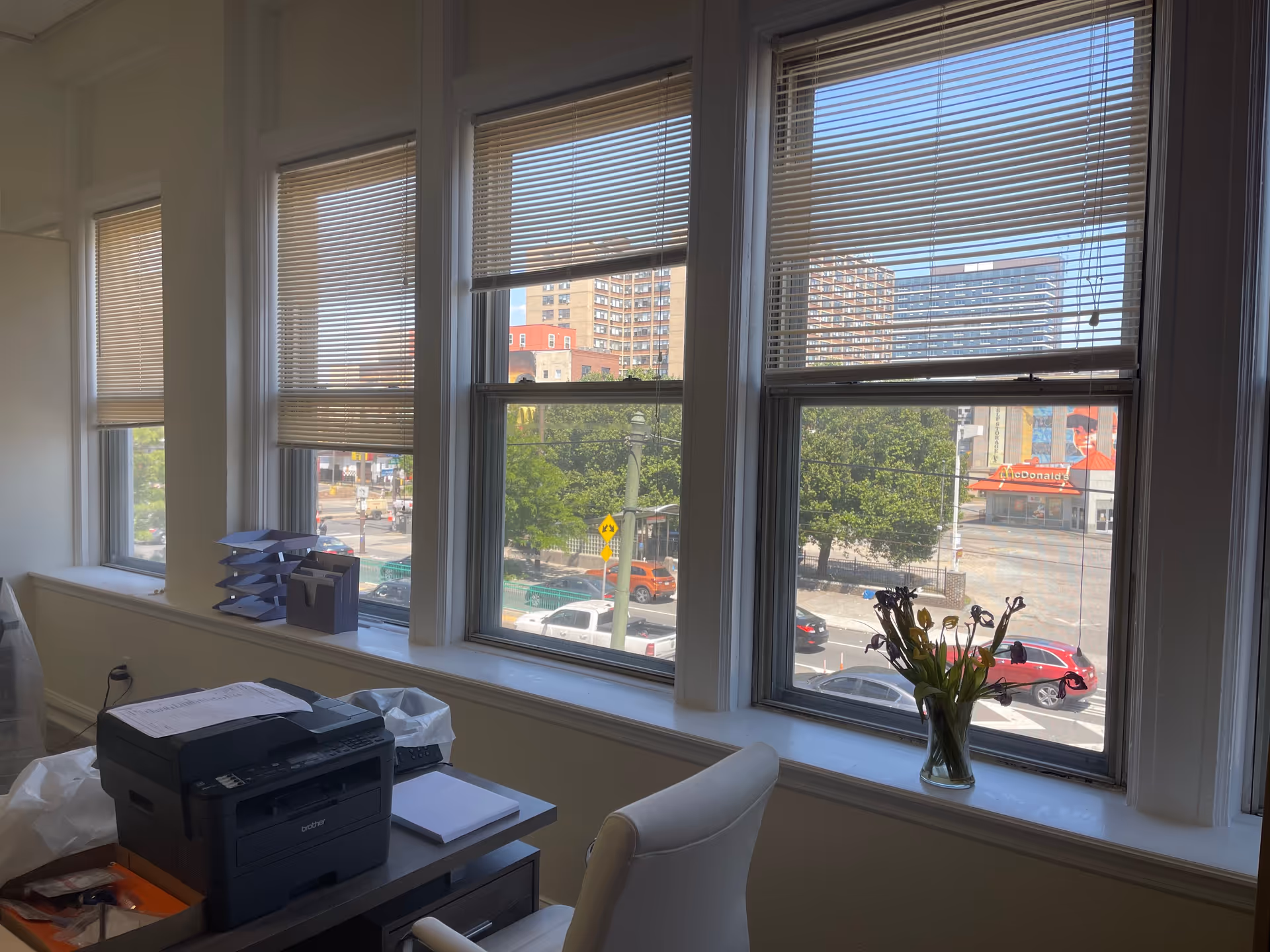 Office corner with printer, paperwork, and a vase on a windowsill overlooking city streets.