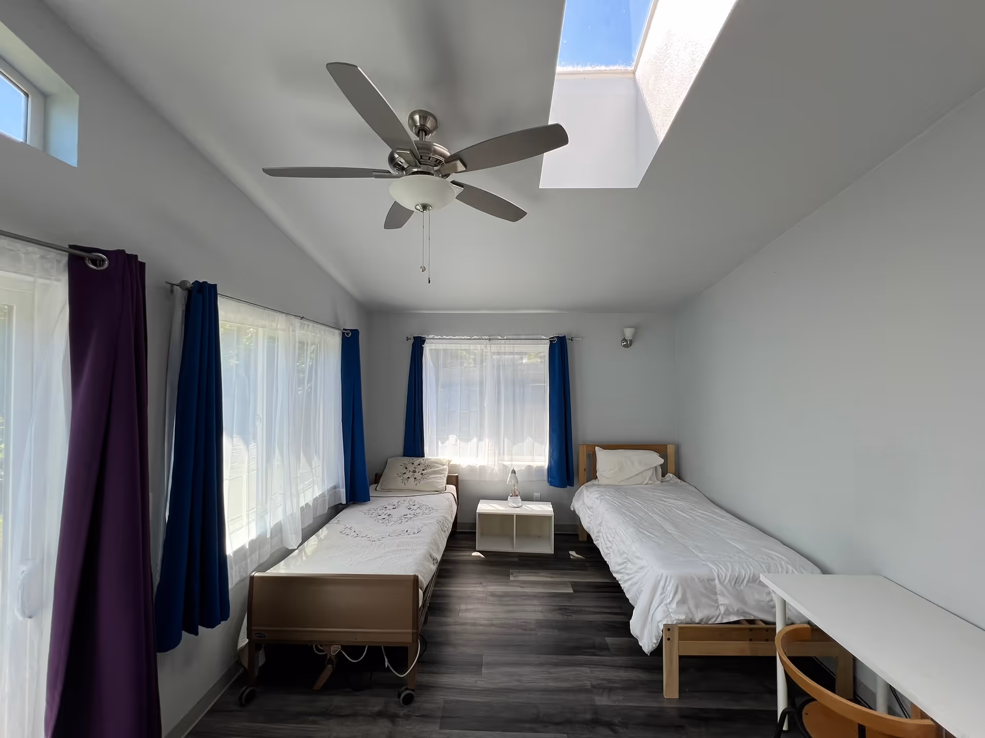 Bright shared bedroom with two single beds, ceiling fan, skylight, and windows.