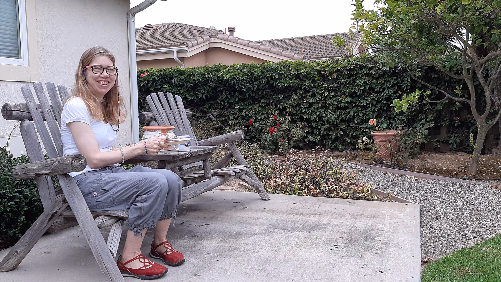 A smiling woman sits on a wooden outdoor bench, holding a teacup.