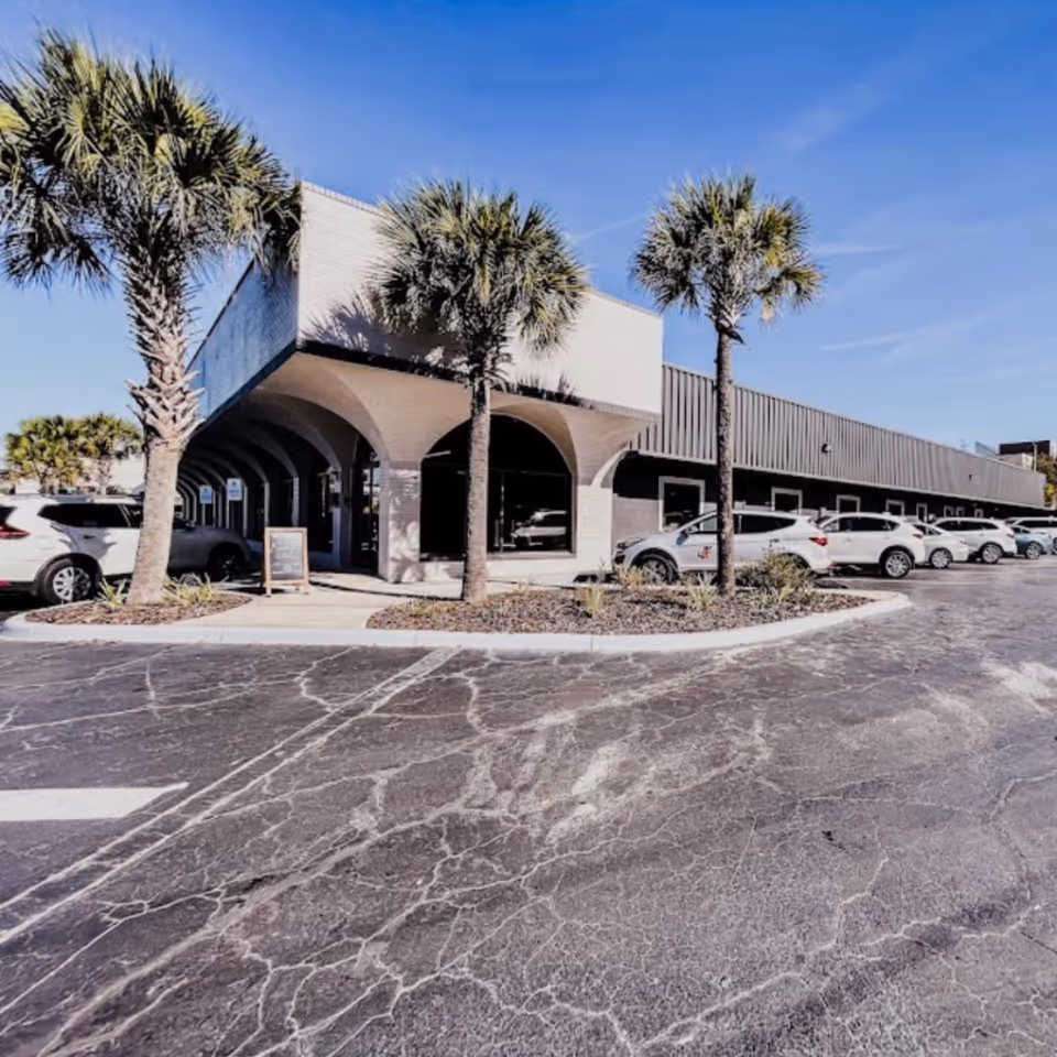 Exterior view of a single-story building with arched entryways, palm trees, and parked cars.