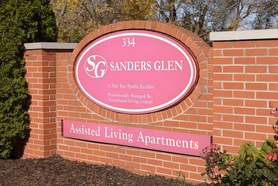 A brick sign for Sanders Glen Assisted Living Apartments with the text '334 SG Sanders Glen A Not For Profit Facility Professionally Managed By, Exceptional Living Centers' on a pink oval background, surrounded by greenery and flowers.