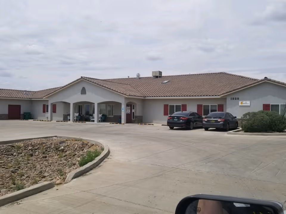 Exterior view of a single-story building with a tiled roof and white walls, featuring red window shutters and a covered entrance area. Two cars are parked in front of the building on a concrete driveway, with a small landscaped area containing rocks and some greenery visible in the foreground under a cloudy sky.
