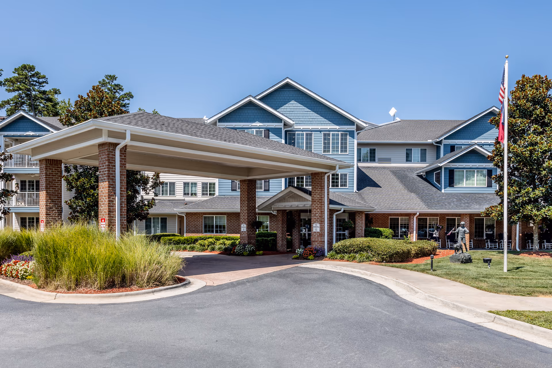 Front entrance of Holiday Cedar Ridge senior living building with a covered porte-cochère, landscaped driveway and flagpole.