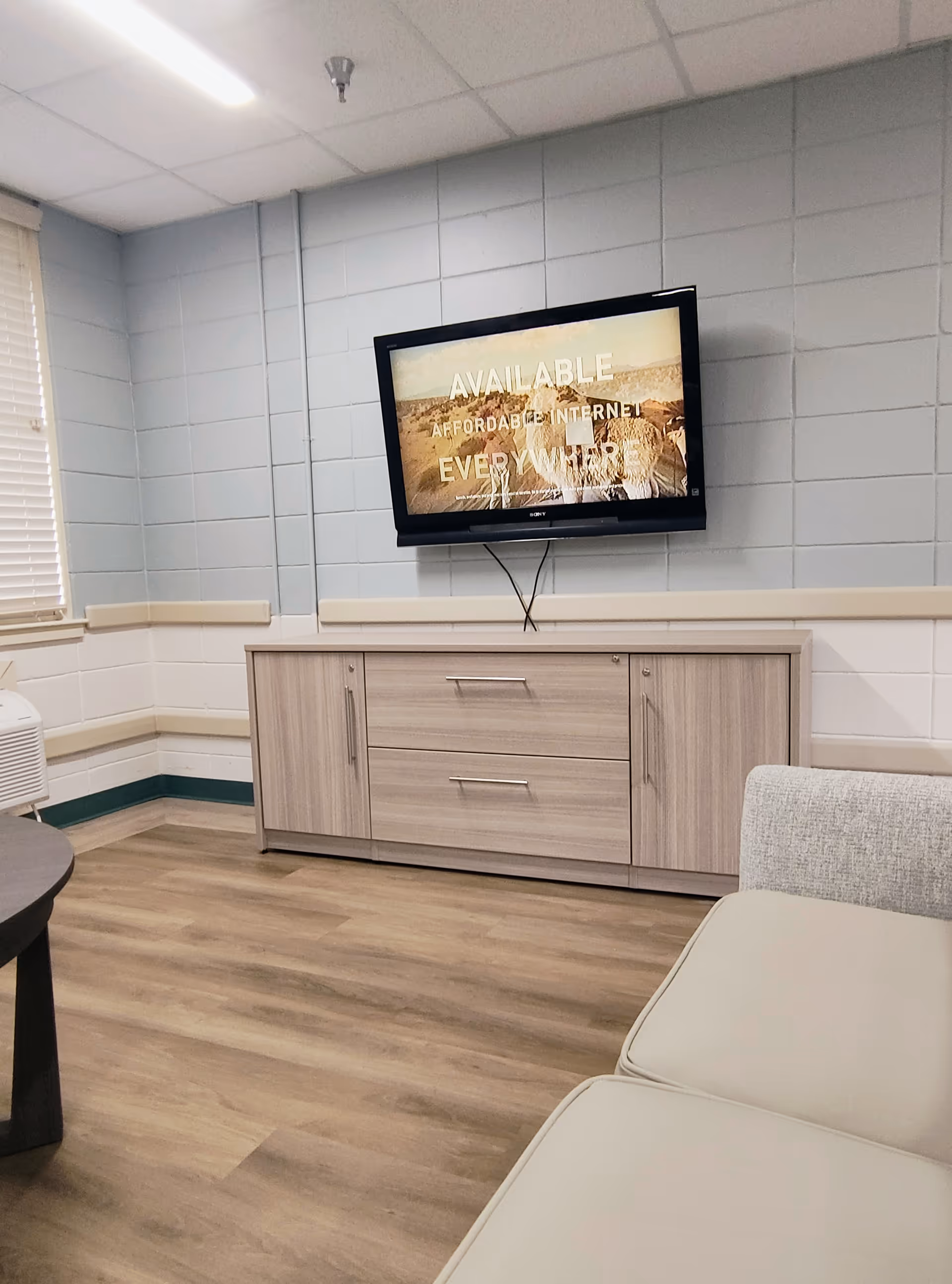 Lounge area with a wall-mounted TV, cabinet, sofa, and wood floor in a senior living facility.