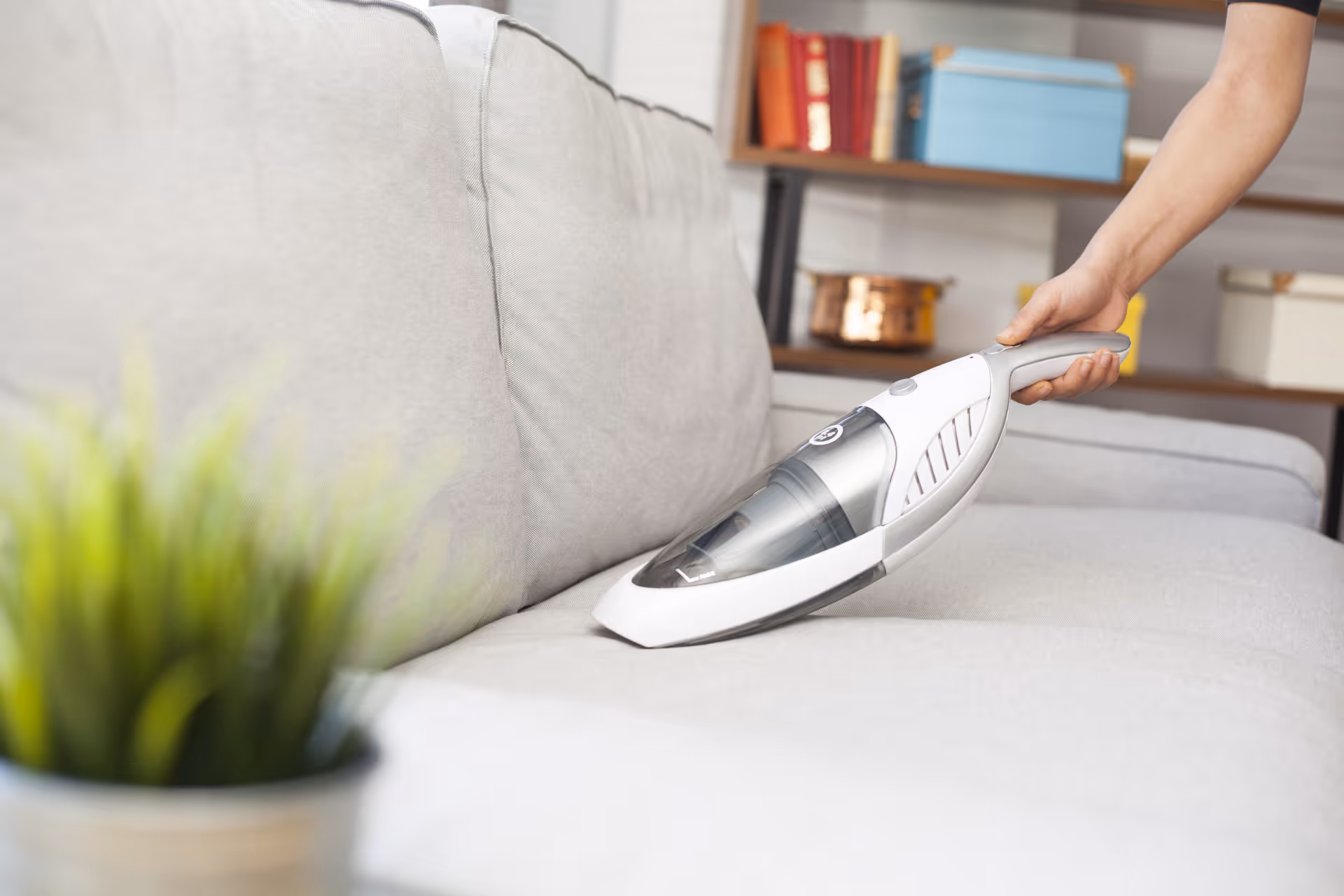 A hand uses a handheld vacuum on a light sofa in a living room.