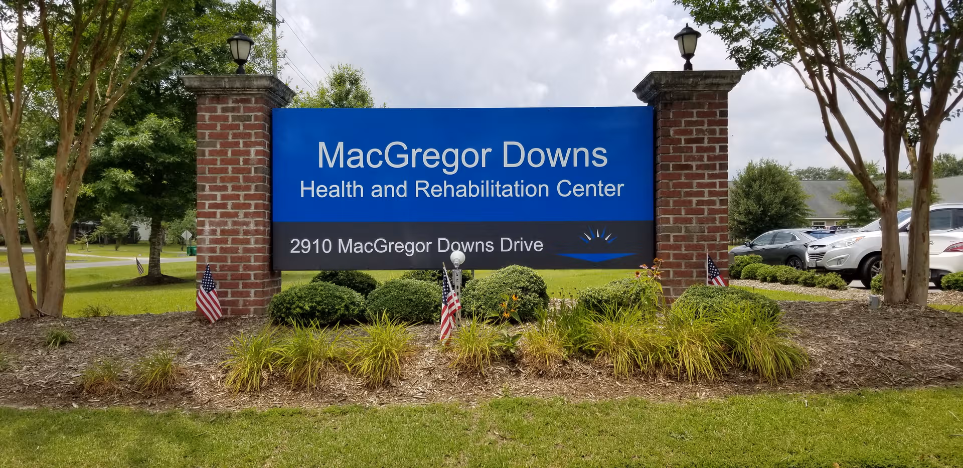 Blue MacGregor Downs Health and Rehabilitation Center sign between brick pillars with landscaping and parked cars.