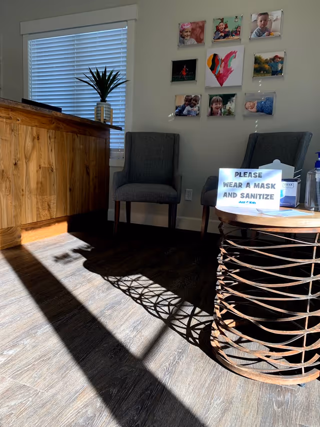 Reception area featuring two gray chairs, wall photos, a plant, and a mask sign.