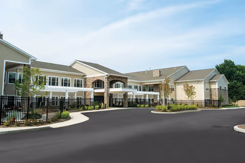 Exterior view of a modern two-story senior living facility with beige siding and stone accents, featuring a covered entrance, landscaped greenery, and a paved driveway under a partly cloudy sky.