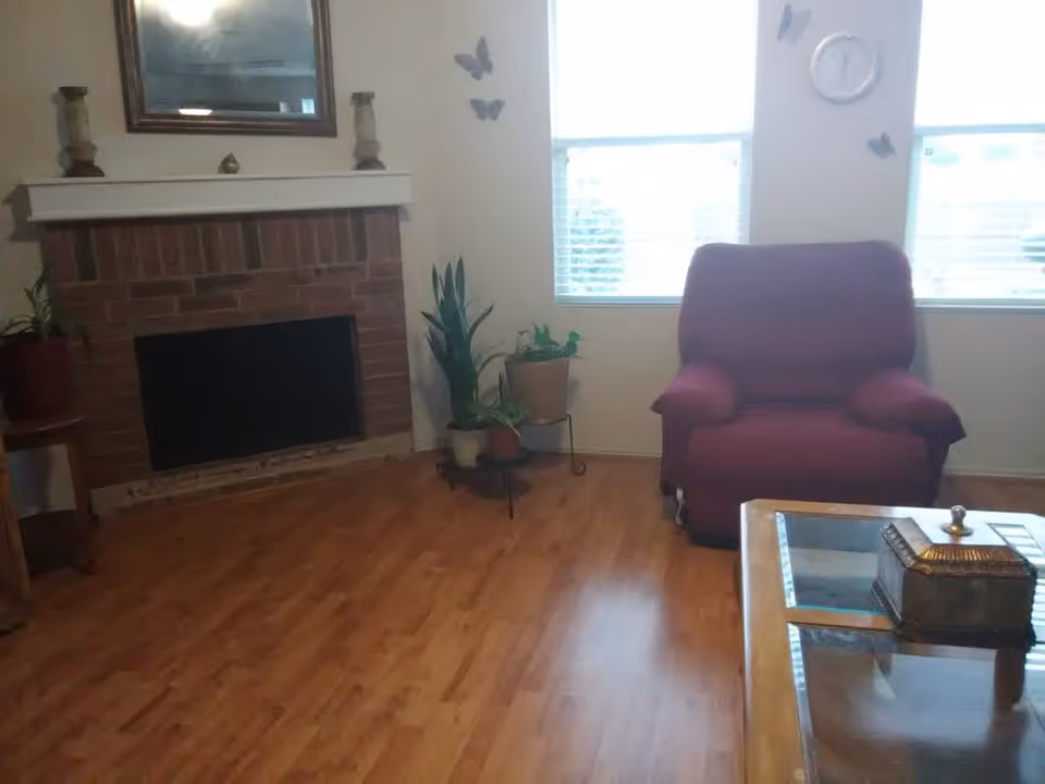 Cozy living room scene with brick fireplace, red armchair, plants, and glass coffee table.