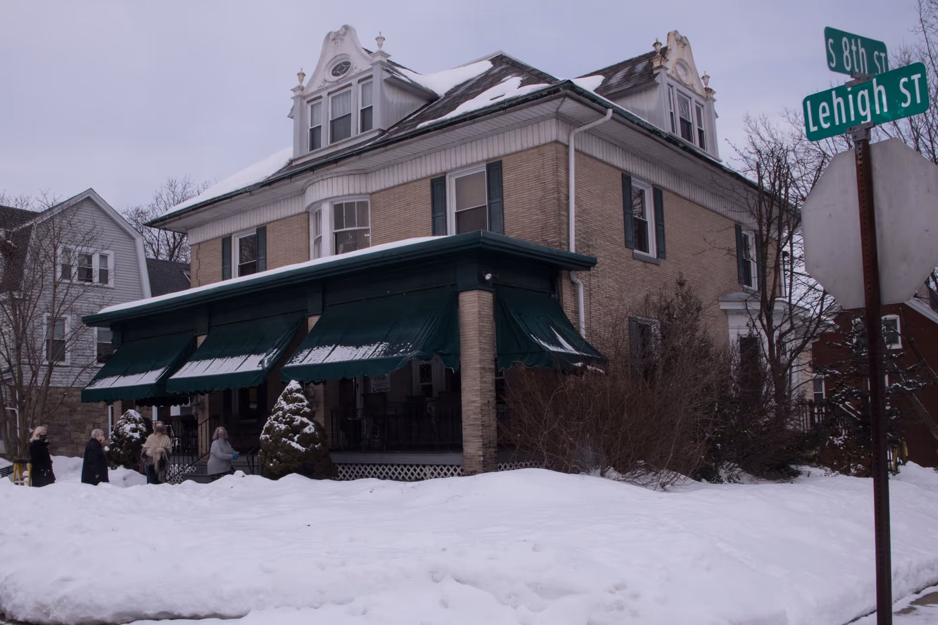 Two-story brick senior living building with green awnings, people outside, and snow on the ground.