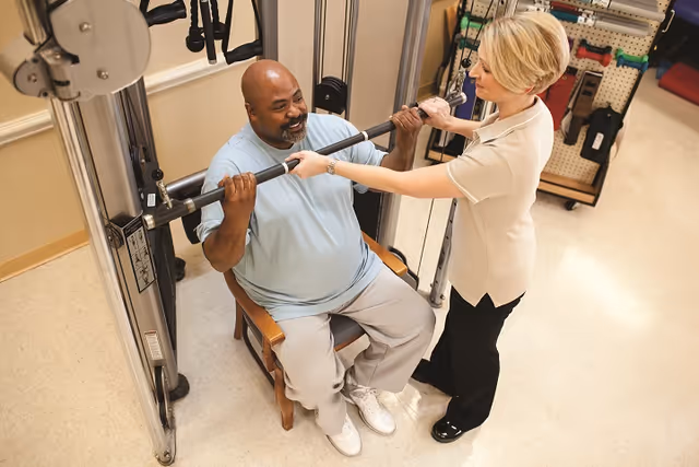 A man seated on a wooden chair is using a weight machine for exercise while a woman stands beside him, assisting and supporting him during the workout in a rehabilitation or fitness room.