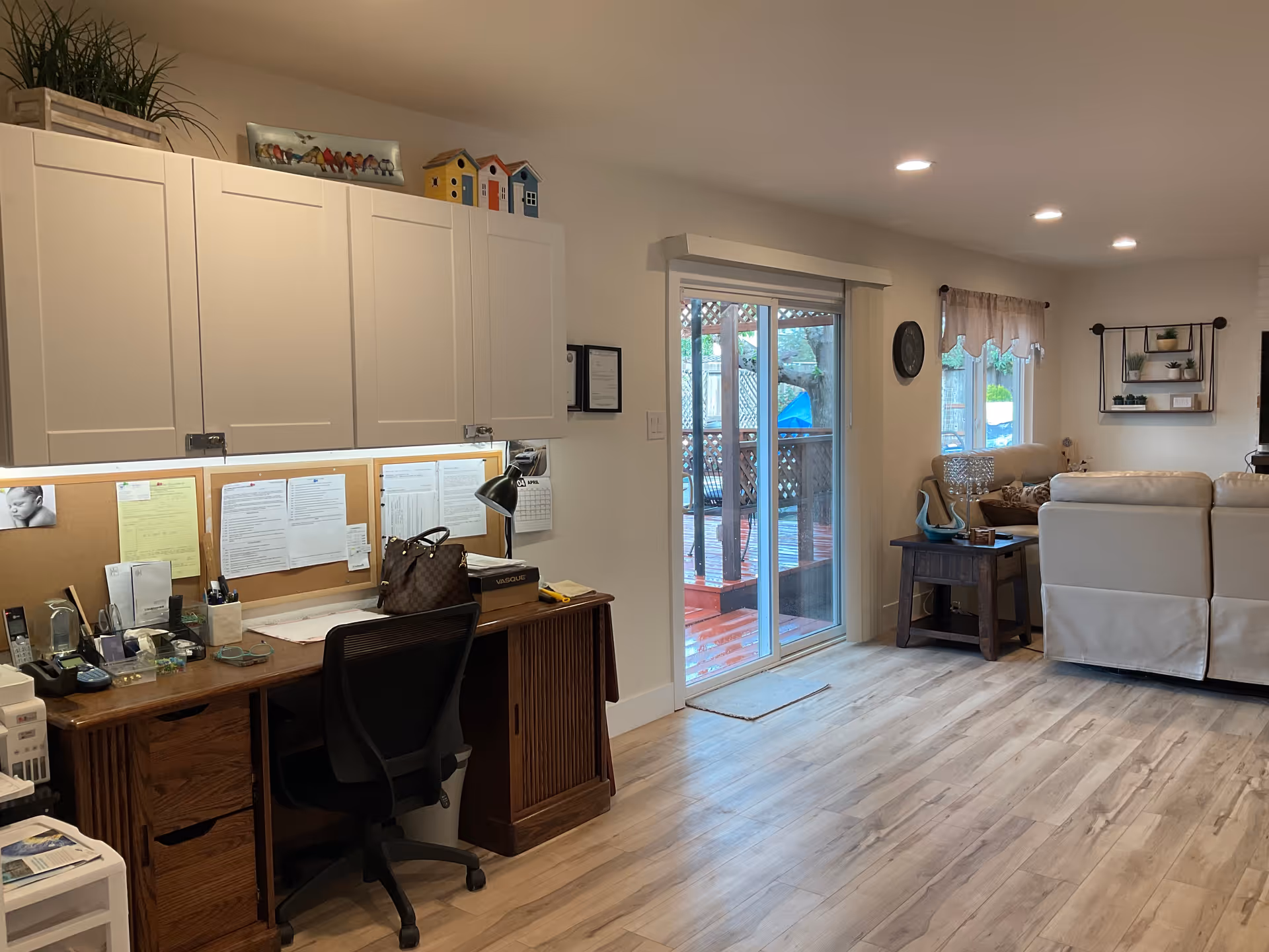 Interior view of a senior living facility room with a wooden desk and office chair on the left, white cabinets above the desk, and a bulletin board with papers pinned on it. To the right, there is a sliding glass door leading outside, a small side table with decorative items, and a beige sectional sofa with a window behind it. The room has light wood flooring and recessed ceiling lights.