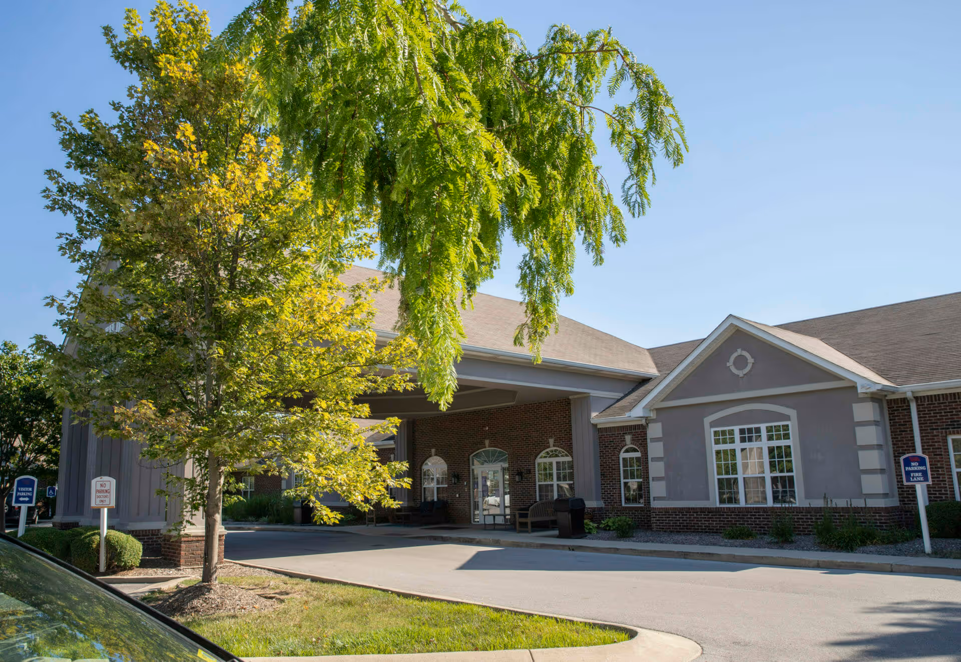 Exterior view of Brownsburg Meadows facility showing a covered entrance with brick and gray siding, surrounded by green trees and a clear blue sky.
