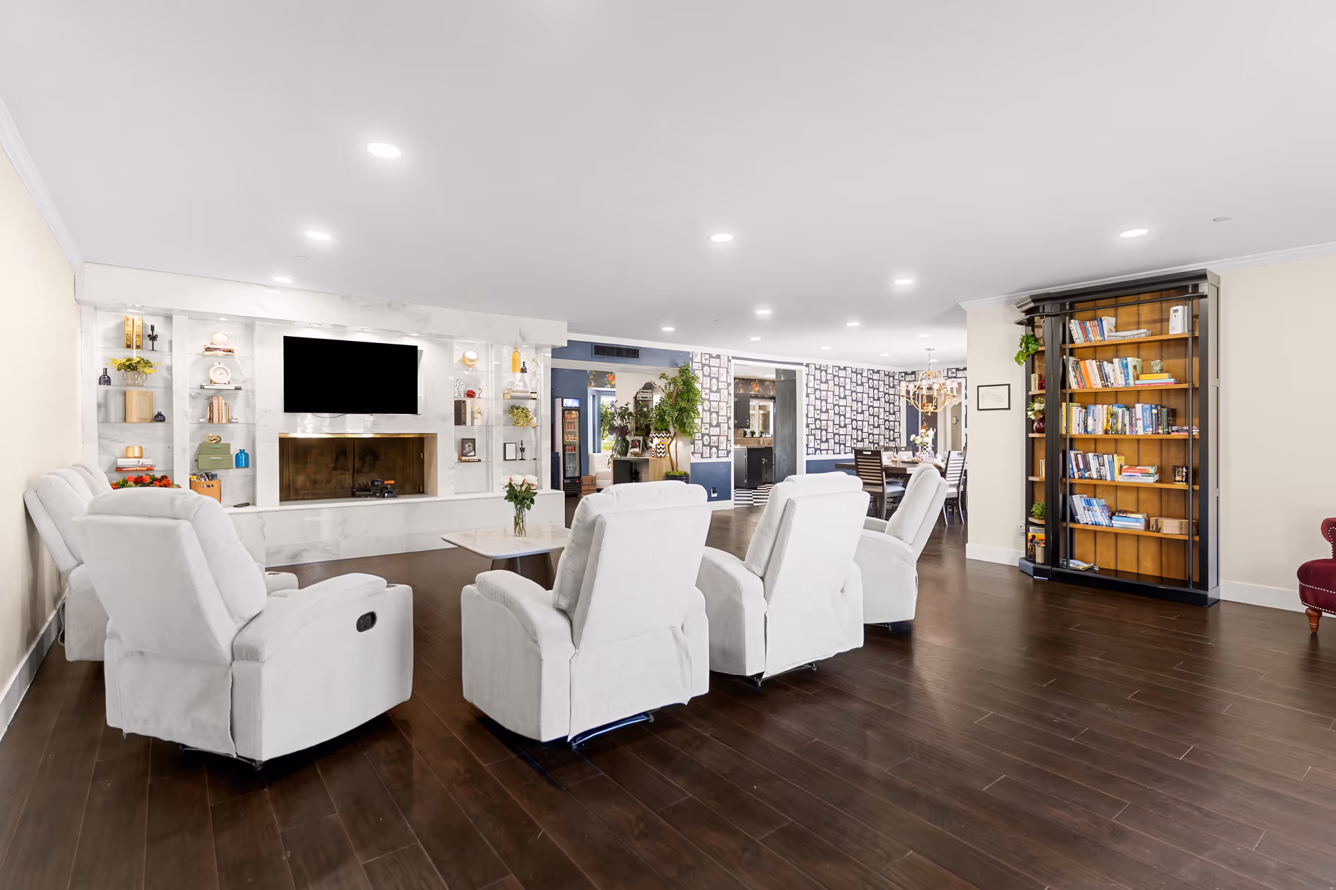 Spacious senior living lounge with white recliners, fireplace, TV, and bookshelves.