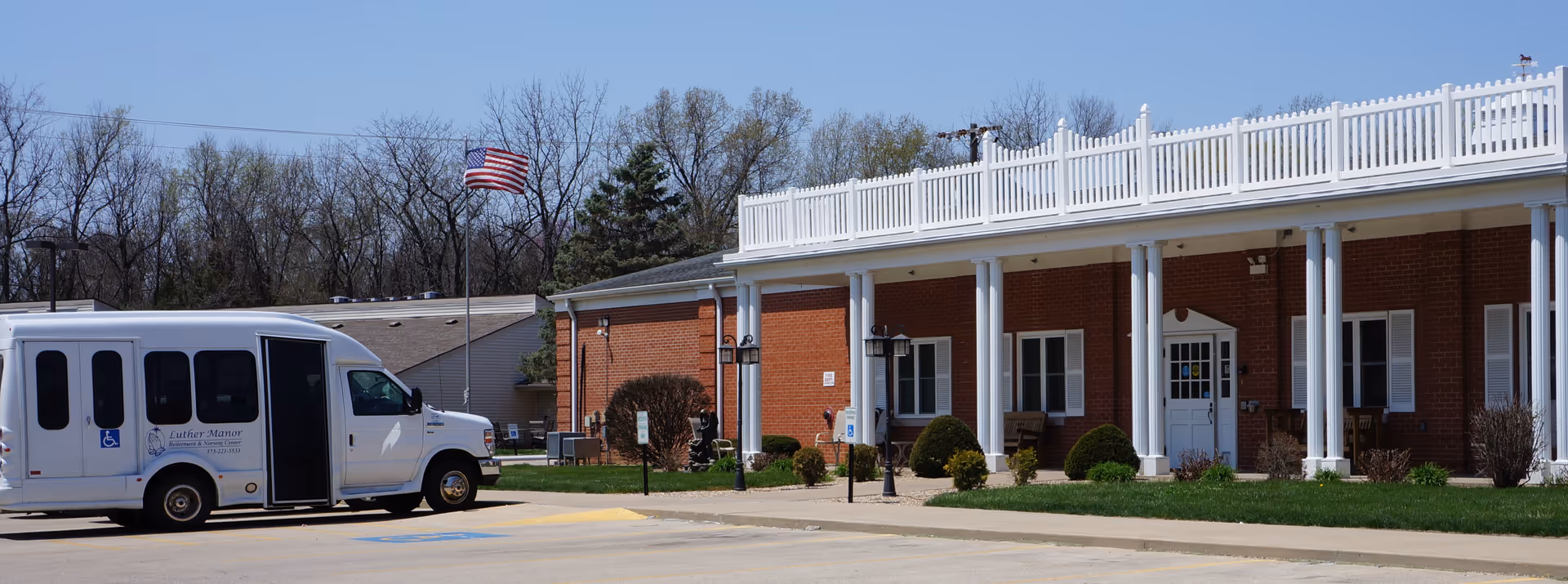 Front entrance of a brick senior living building with white columns, a parked shuttle van, and an American flag.