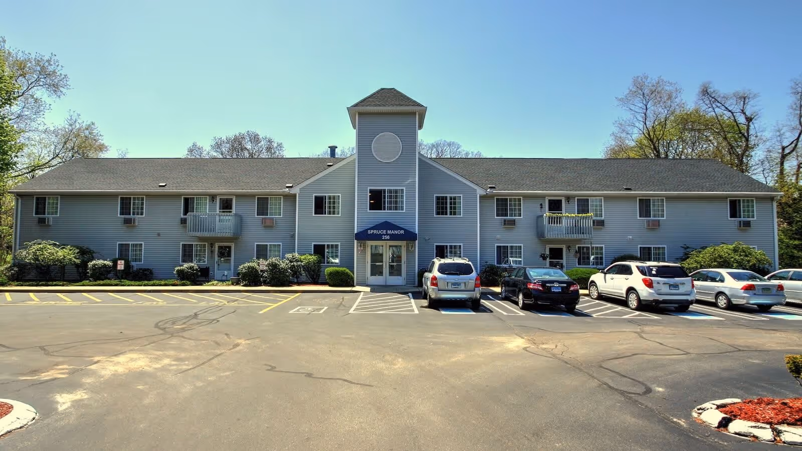 Front exterior view of Spruce Manor, a two-story building with gray siding, multiple windows, small balconies, and a central entrance with a blue awning displaying the facility name and number 256. Several cars are parked in front of the building in designated parking spaces, including handicapped spots. Trees and shrubs surround the building under a clear blue sky.