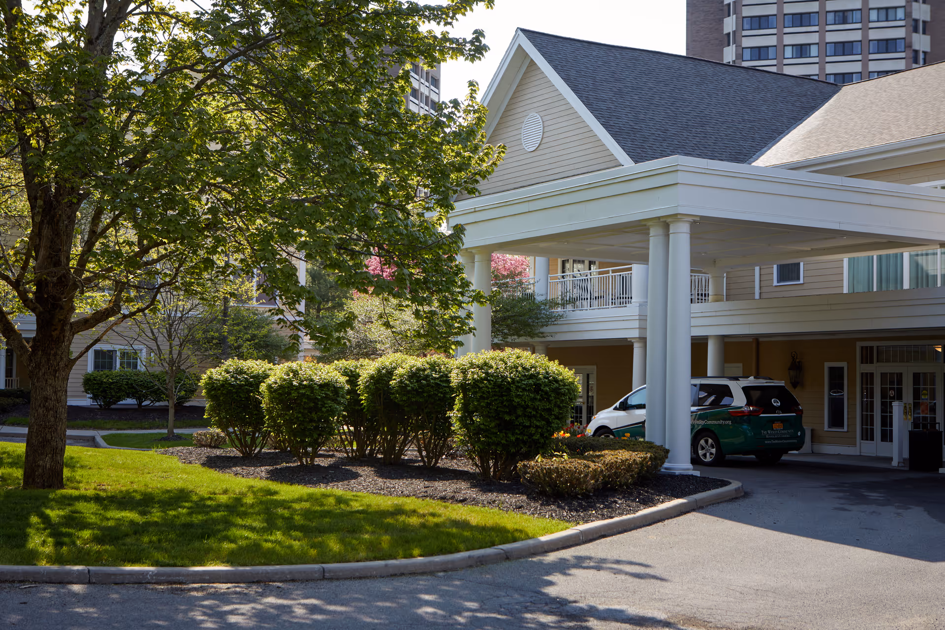 Covered entrance/portico of a senior living building with landscaped shrubs, a tree, and a parked van.