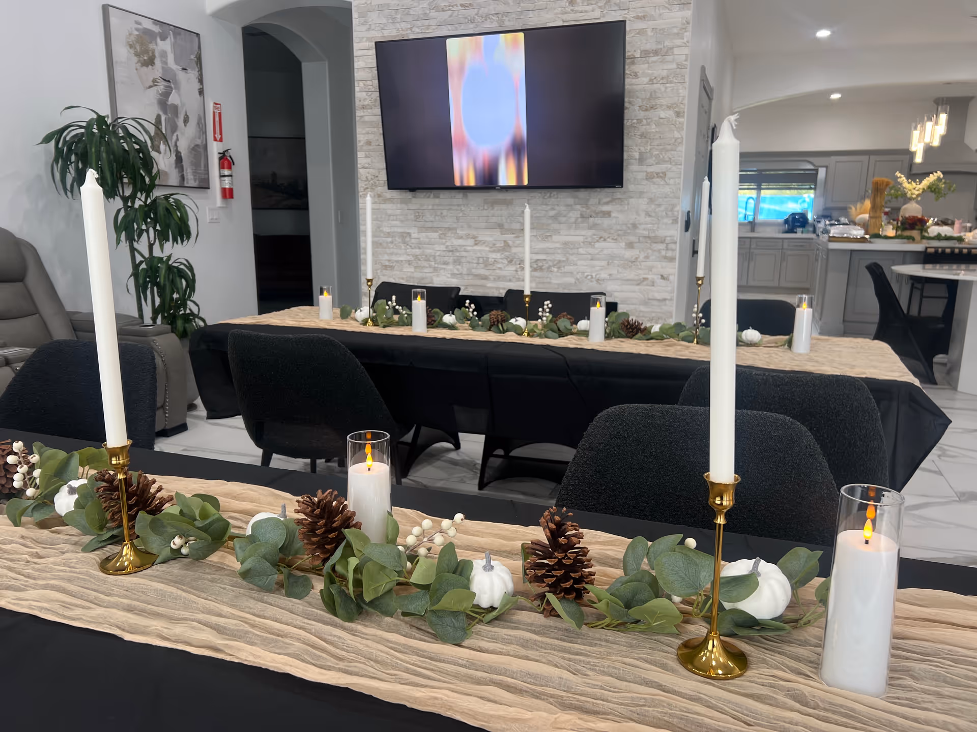 Long dining table with candles, pinecones, greenery, and a TV mounted on a stone feature wall.
