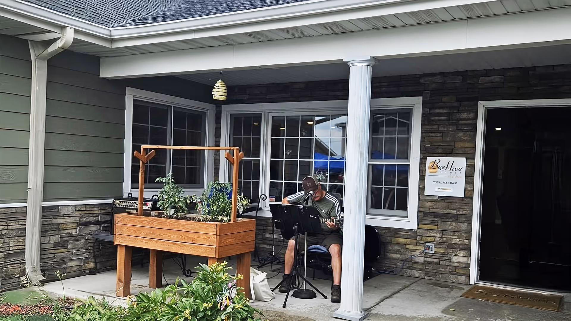 Resident playing guitar on a covered outdoor porch beside a planter and BeeHive Homes sign.