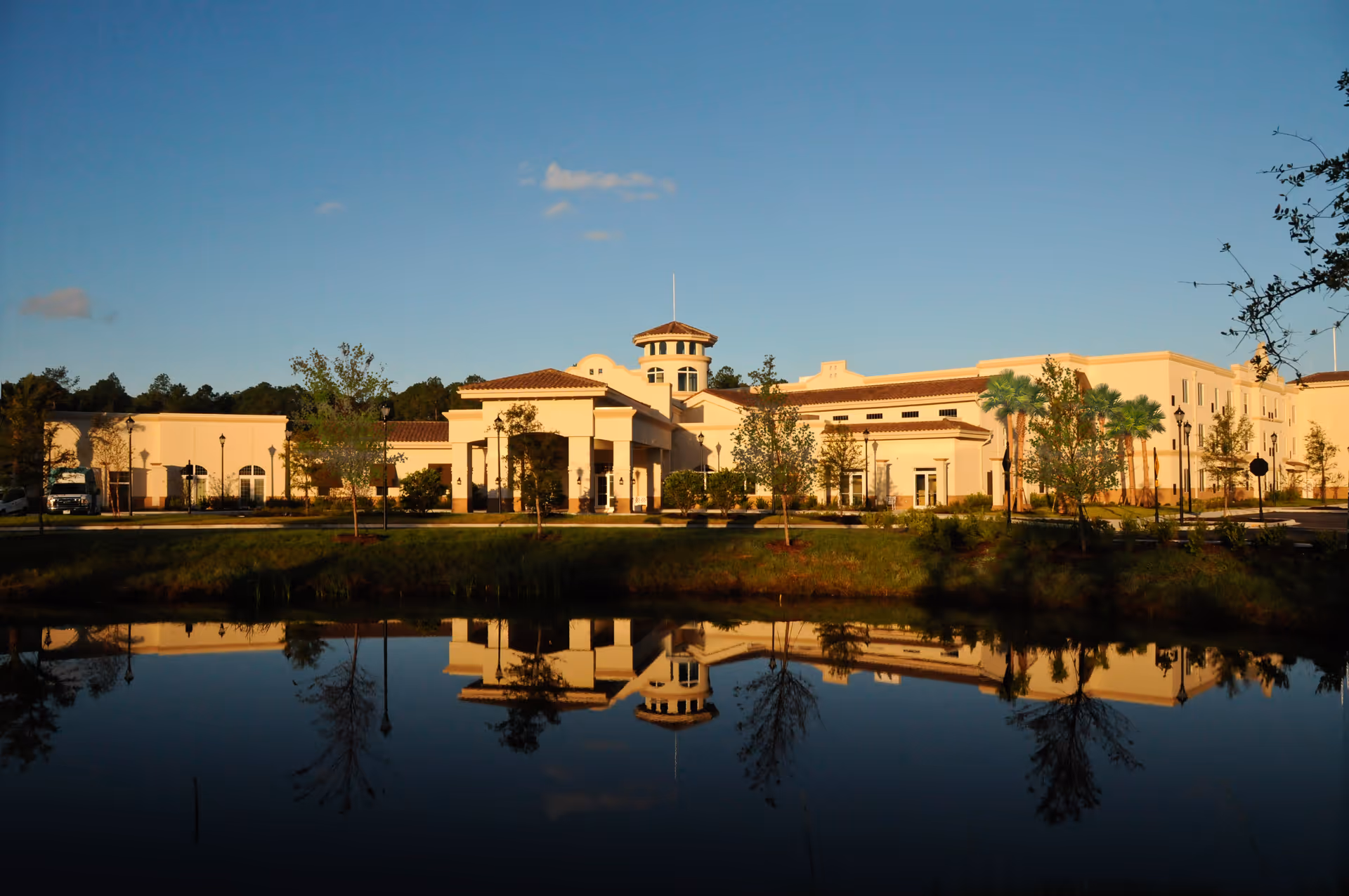 Exterior view of a large, beige senior living facility named Silver Creek, with a tiled roof and a central tower. The building is reflected in a calm pond in the foreground, surrounded by trees and greenery under a clear blue sky.