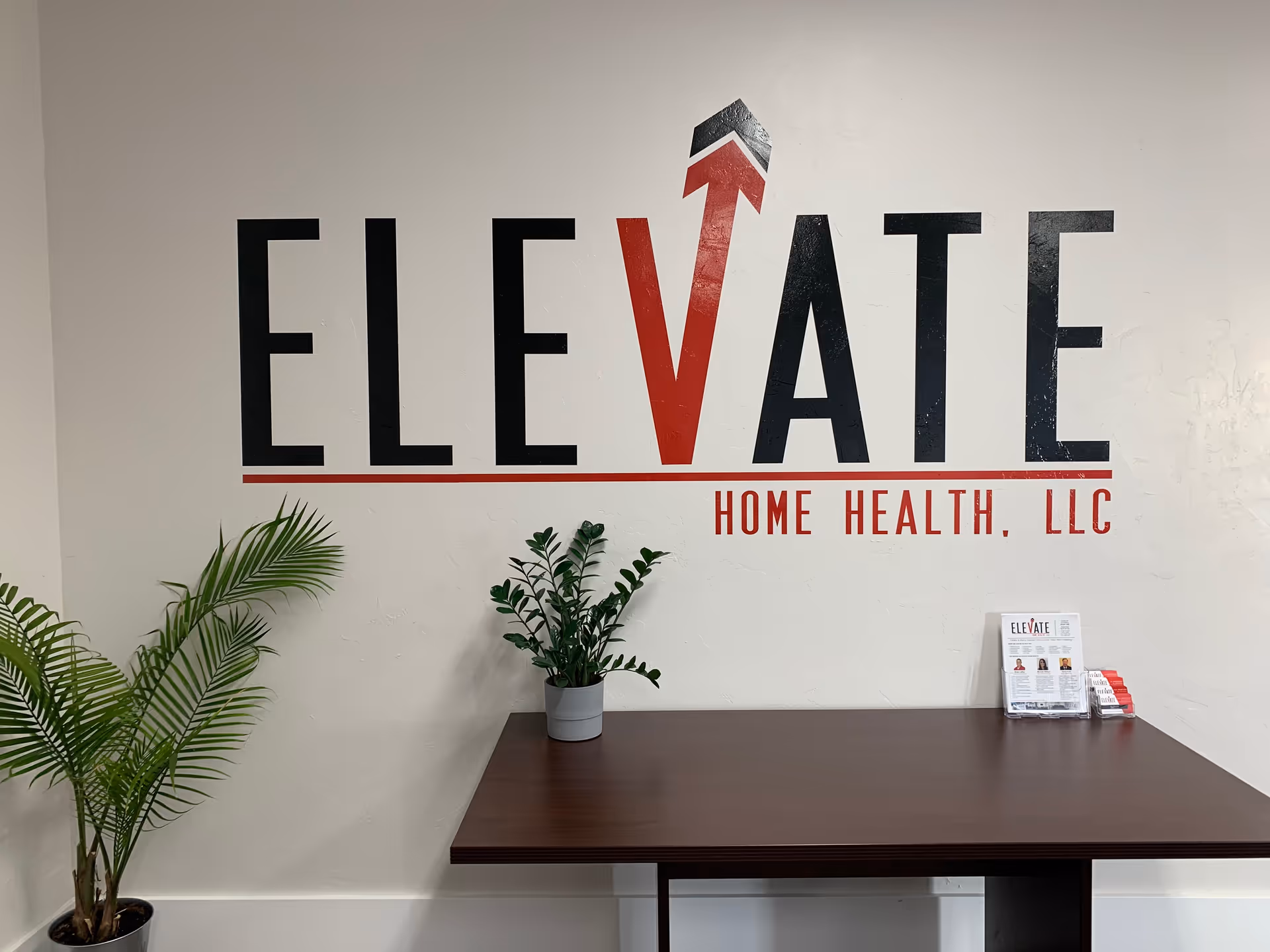 Interior wall featuring Elevate Home Health, LLC logo, a wooden table, and potted plants.