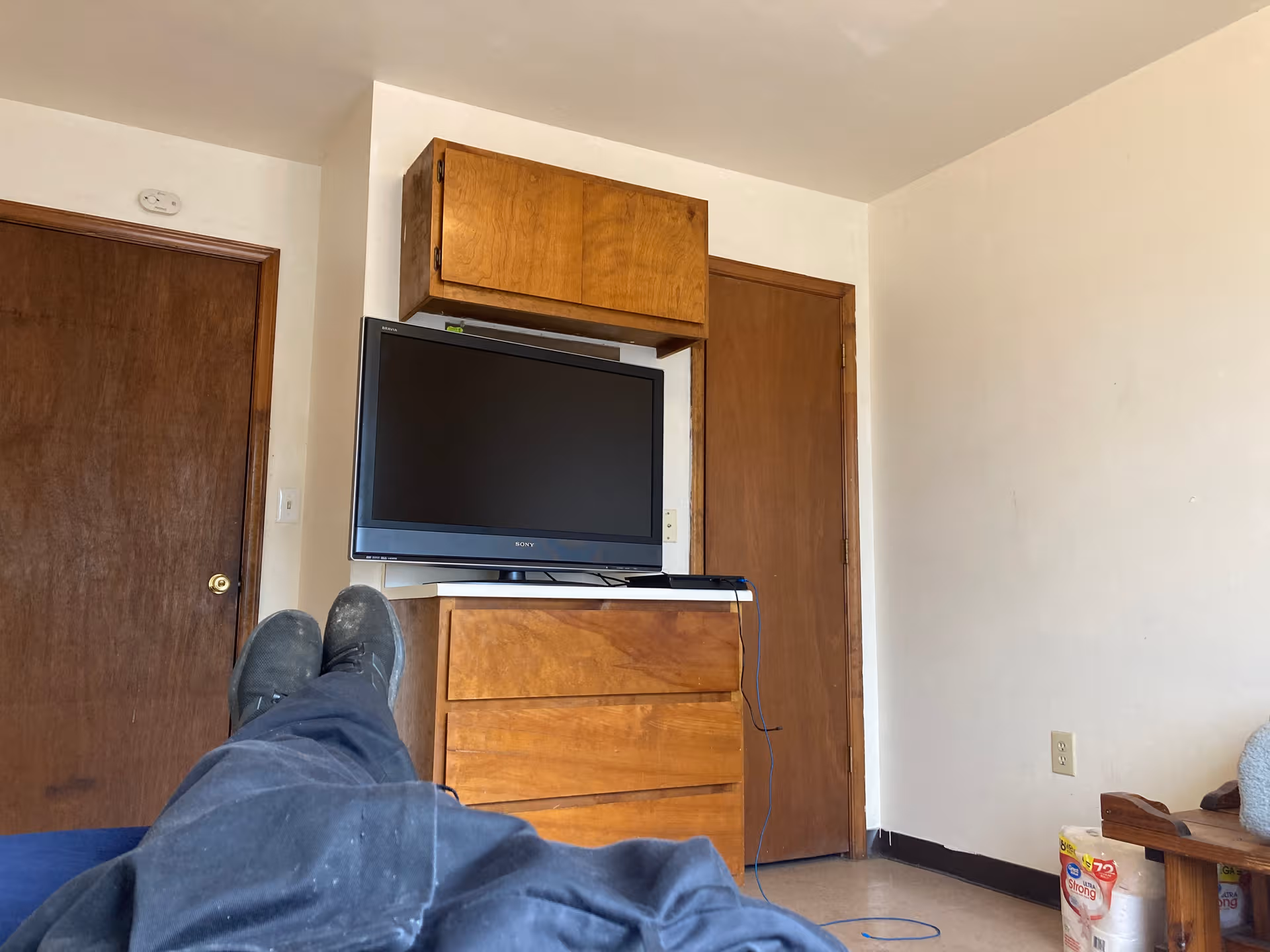 Resident's legs visible in bed facing a TV atop a dresser in a small, plain room.