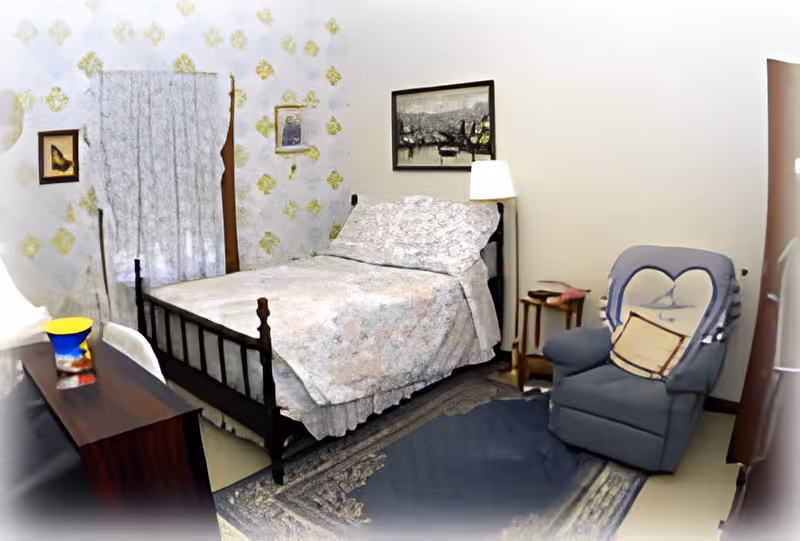A cozy bedroom with a bed covered in a floral bedspread, a blue armchair with a heart-shaped pillow, a wooden side table with a lamp, a dresser with a colorful vase, and framed pictures on the walls. The room has patterned wallpaper and a curtain covering a window.