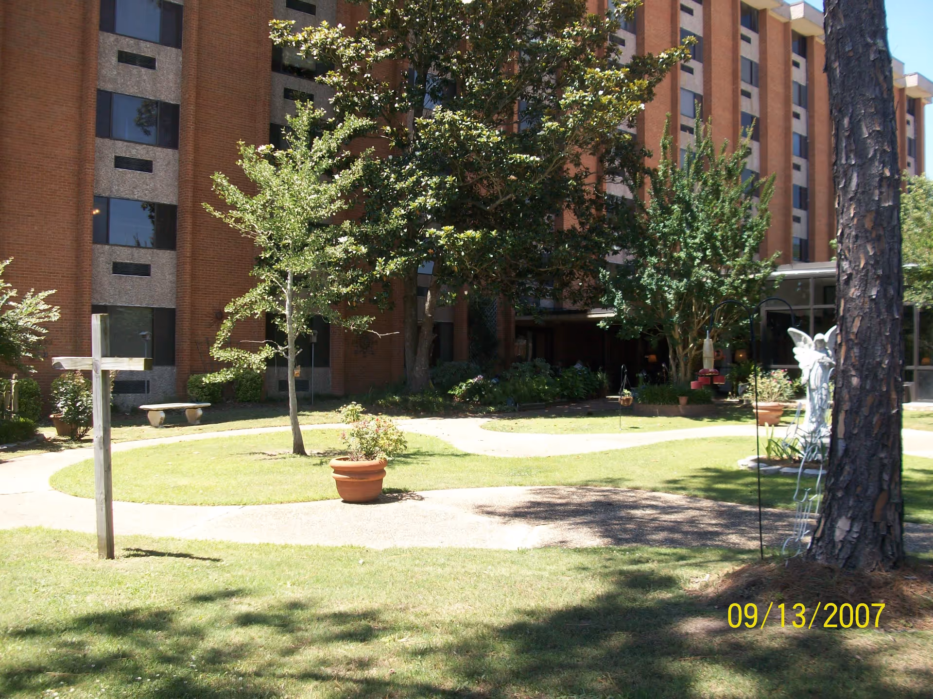 Brick retirement community building with a grassy courtyard, winding paths, potted plants and trees.