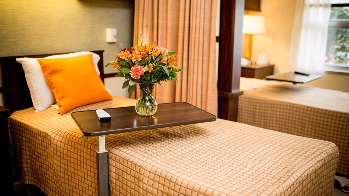 Two twin beds in a senior living bedroom with an overbed table holding a vase of flowers and a remote.